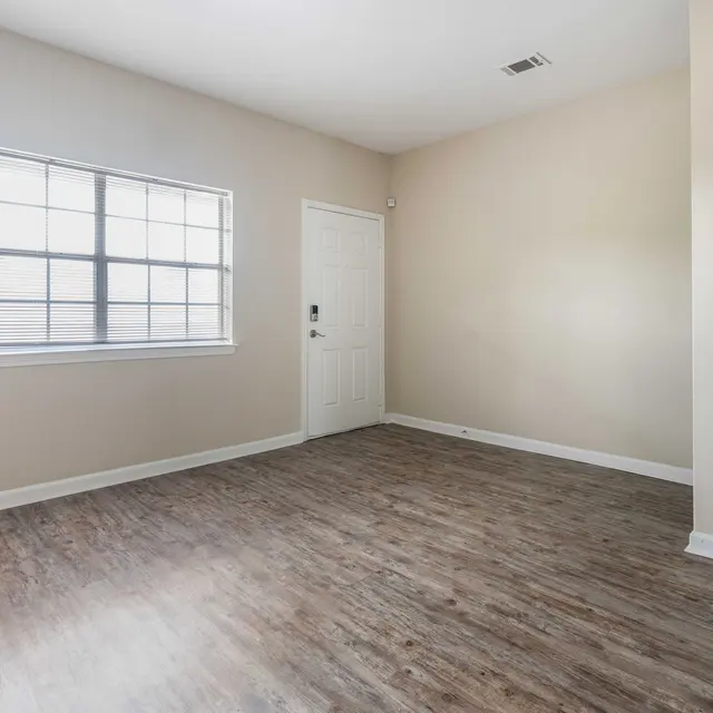 An empty room with light wooden flooring, beige walls, and a window on the left side. A door is visible on the right side.
