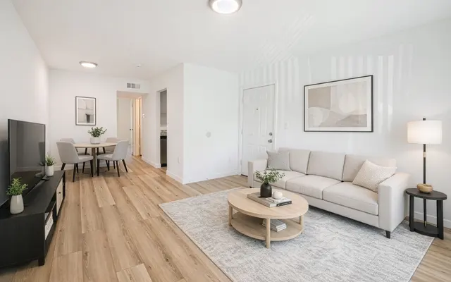 Modern Living Room Interior A bright and modern living room featuring a cozy couch, a circular coffee table, and a TV stand. The floor is made of light wood, and there are decorative plants on furniture. The walls are painted white and have a framed artwork. A dining area is visible in the background.