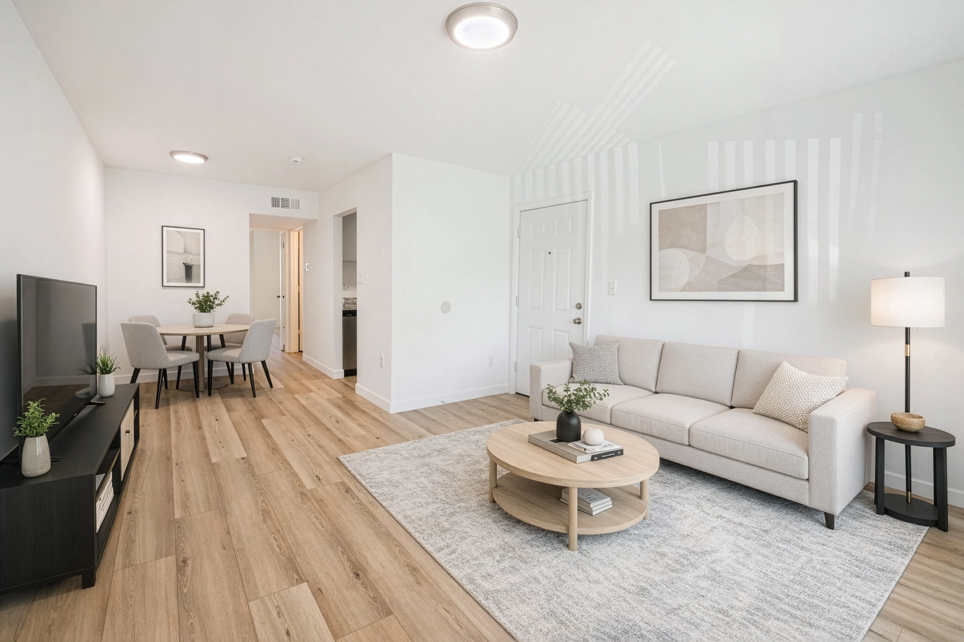 Modern Living Room Interior A bright and modern living room featuring a cozy couch, a circular coffee table, and a TV stand. The floor is made of light wood, and there are decorative plants on furniture. The walls are painted white and have a framed artwork. A dining area is visible in the background.