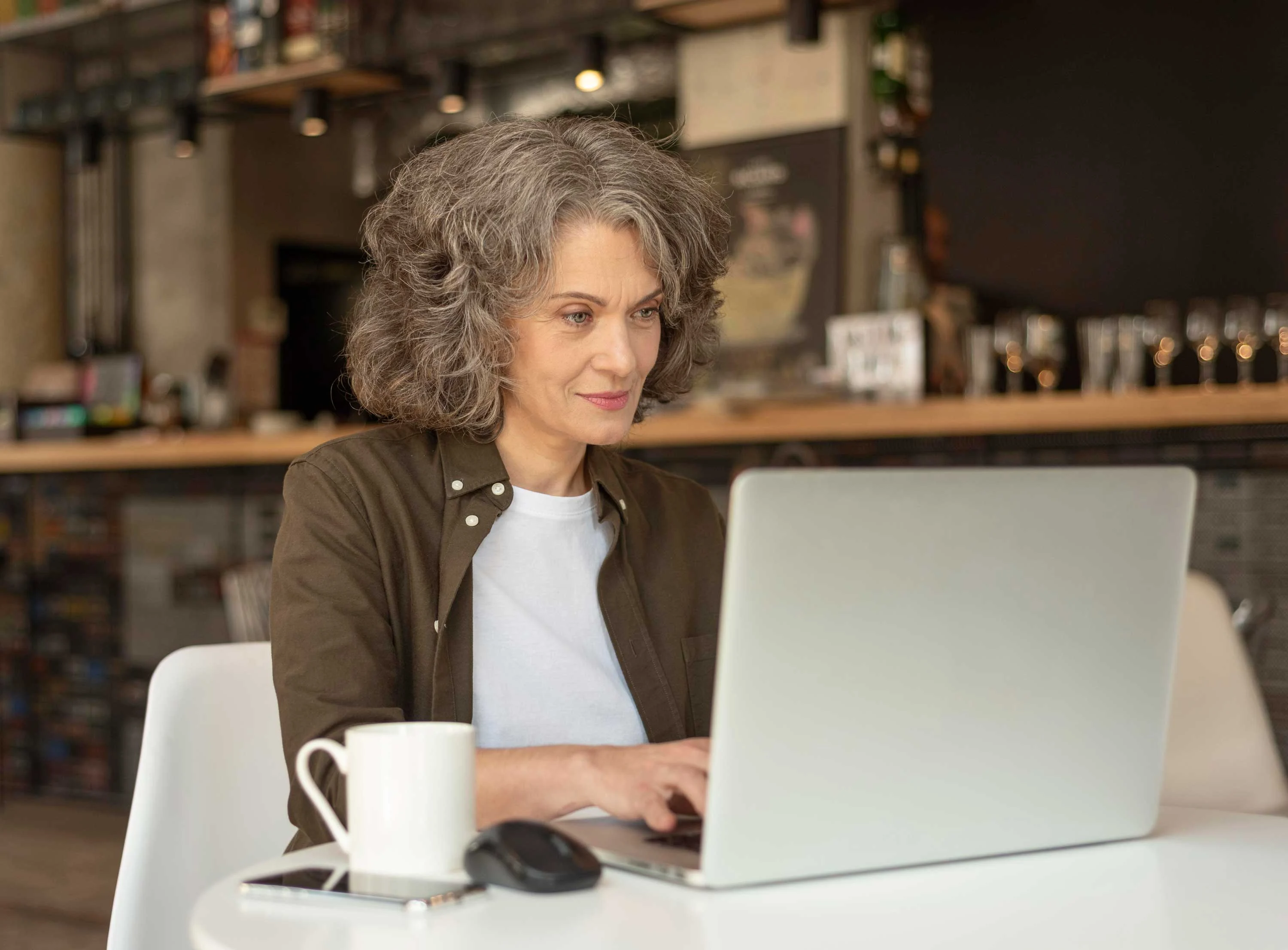 A woman with curly gray hair is sitting at a table in a cafe, working on a laptop. A white cup and a computer mouse are on the table beside her.