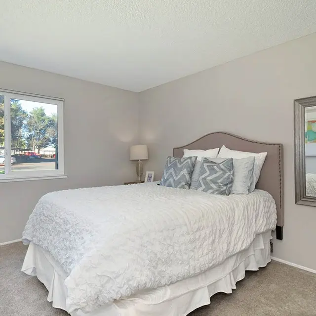 A cozy bedroom featuring a white bed with decorative pillows, a bedside lamp, and a large mirror. There is a window showing a view of greenery outside.