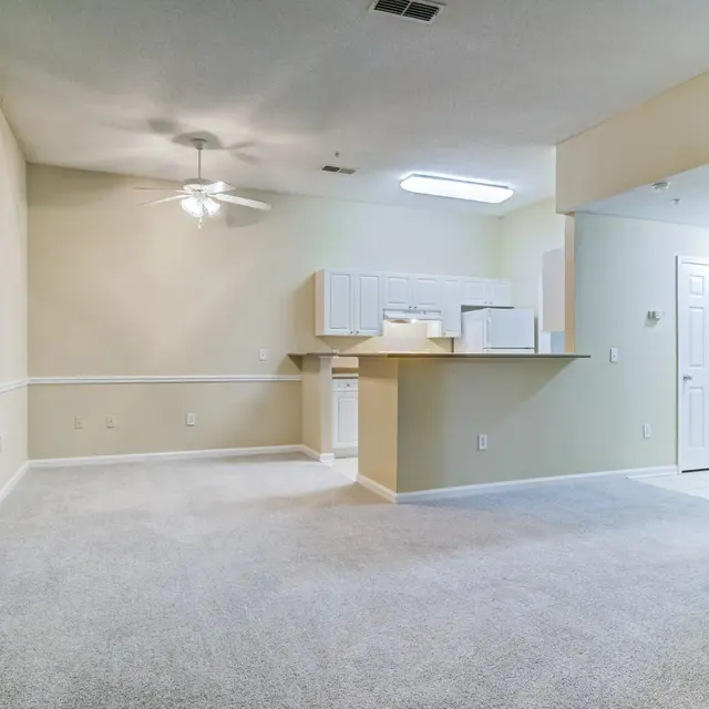 An open-concept living space featuring a beige carpeted floor, a ceiling fan, and a kitchenette with white cabinetry. The walls are painted a light color, and there's a door on the left side leading to another room.