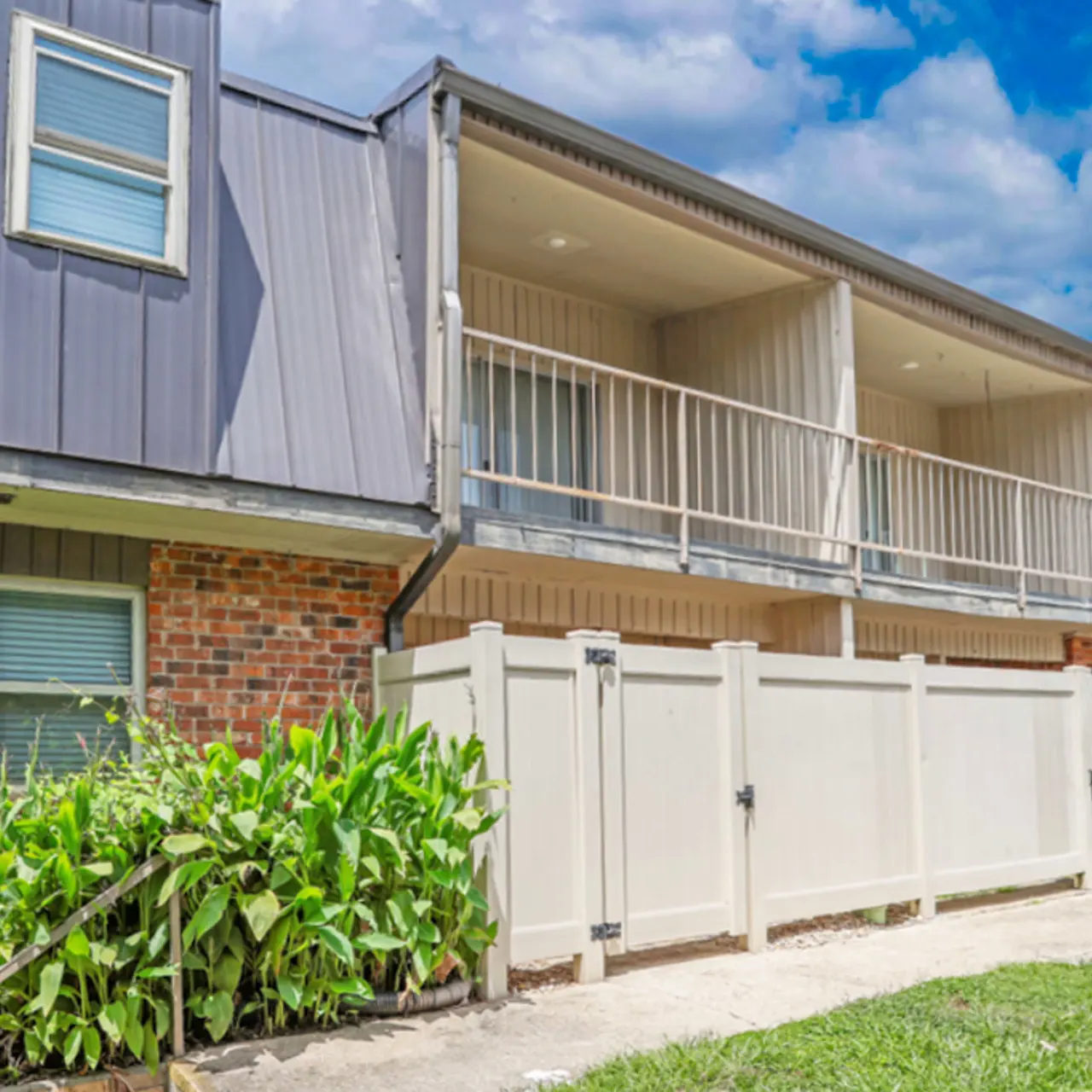 Exterior view of an apartment building featuring a balcony, surrounded by greenery and fencing.