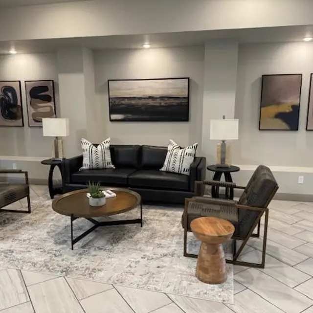 A modern living room featuring a black leather sofa with patterned cushions, two armchairs, and a round coffee table on a decorative rug. There are two lamps on either side of the sofa and abstract art pieces hanging on the wall.