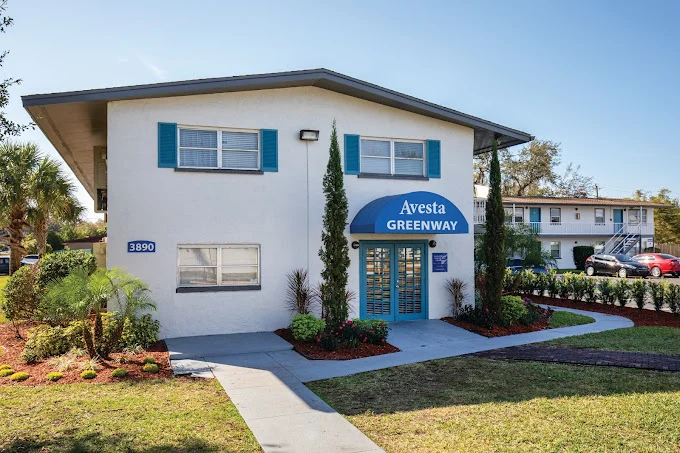 Exterior view of a two-story building with blue accents and a sign reading 'Avesta Greenway.' The entrance is marked by an archway with a blue awning. The surrounding area features landscaped greenery and a parking lot visible in the background.
