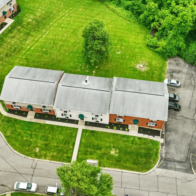 Aerial view of a residential complex with several buildings surrounded by green trees and grass, featuring a circular driveway and parked cars.