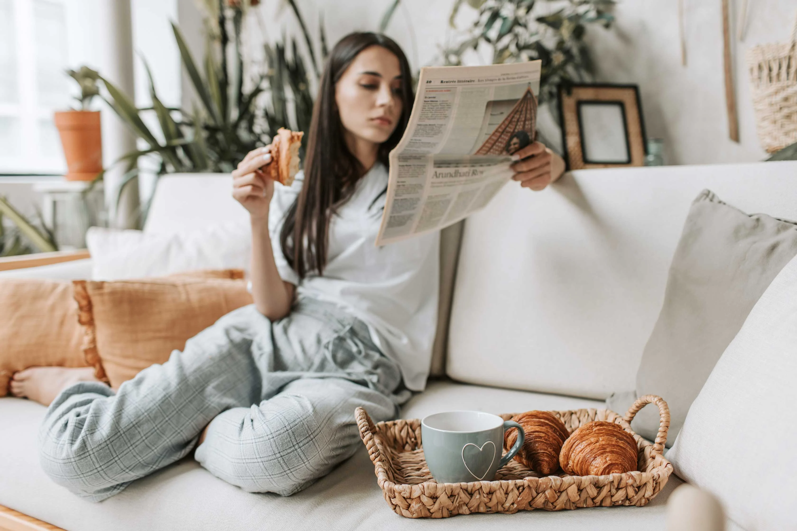 A woman sitting on a couch, enjoying a croissant while reading a newspaper. A tray with a coffee cup and croissants is placed in front of her.