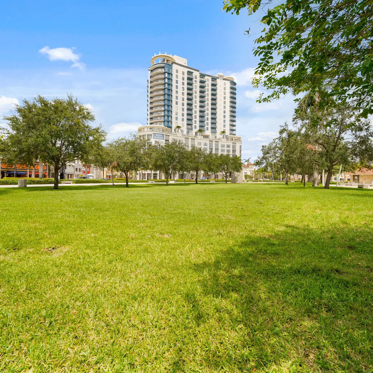 A green park area with grass and trees, featuring a tall building in the background under a clear blue sky.