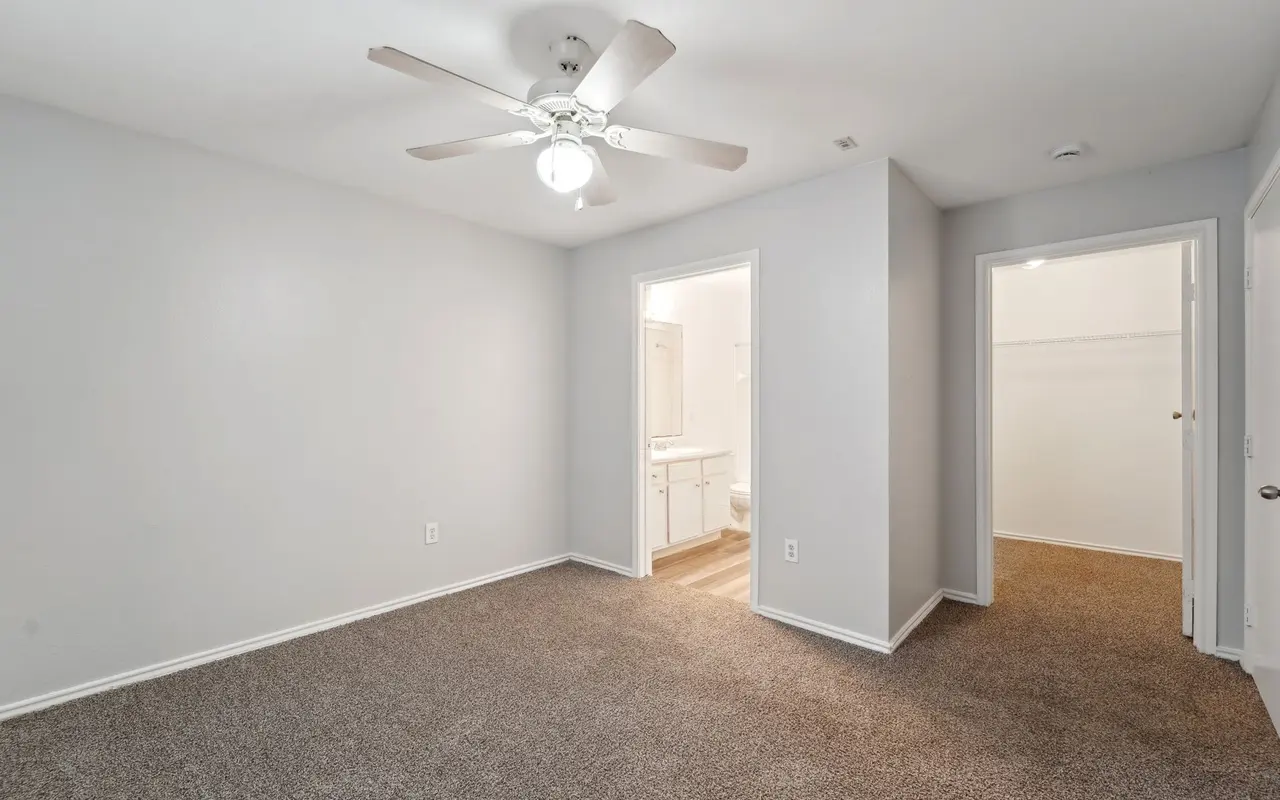 An empty room with light gray walls and carpet, featuring a ceiling fan and two doorways leading to other areas.