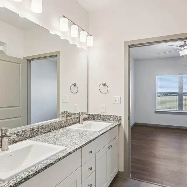 A modern bathroom with a double vanity featuring a granite countertop, two sinks, and a large mirror with bright lighting. A door leads to a room with a ceiling fan and a window.