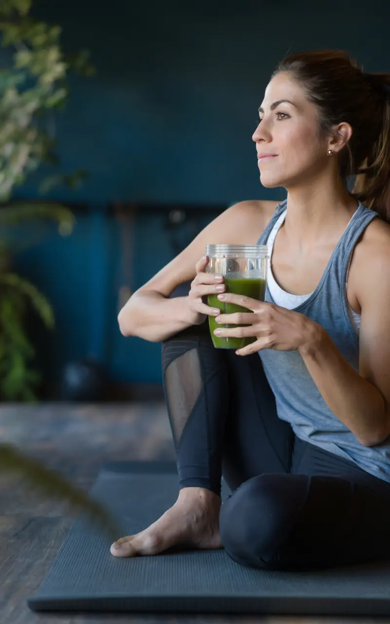 Wellness and Mindfulness in Yoga A woman sits cross-legged on a yoga mat, holding a green beverage, with a serene expression contemplatively gazing outside. The background features indoor plants and a peaceful, well-lit space.