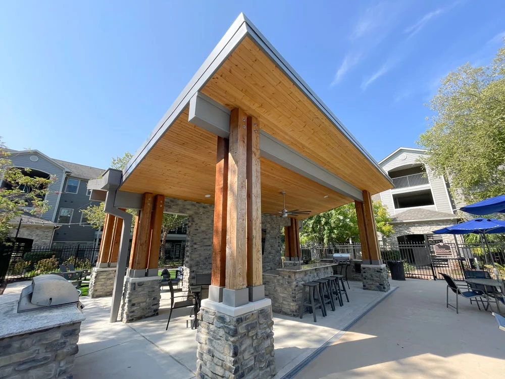 Outdoor Pavilion Area An outdoor pavilion area featuring wooden beams, stone columns, and a shaded seating area. The pavilion is located near a pool and there are grill facilities visible in the foreground.