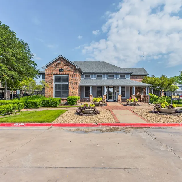 A brick building with large windows surrounded by landscaped greenery and flower beds under a partly cloudy sky.