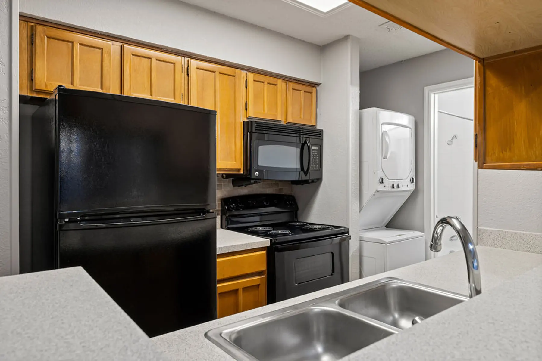 Modern Kitchen Design A modern kitchen featuring a black refrigerator, black microwave, and black stove, with wooden cabinets and a double sink in the foreground. A stacked washer and dryer are located in the corner of the kitchen.