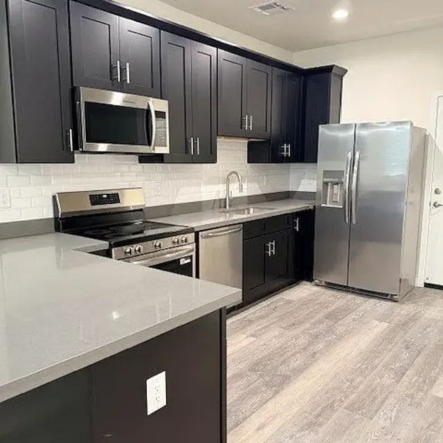 A modern kitchen featuring dark wooden cabinets, stainless steel appliances, and a large countertop. The kitchen has a sleek and contemporary design with gray countertops and light-colored flooring.
