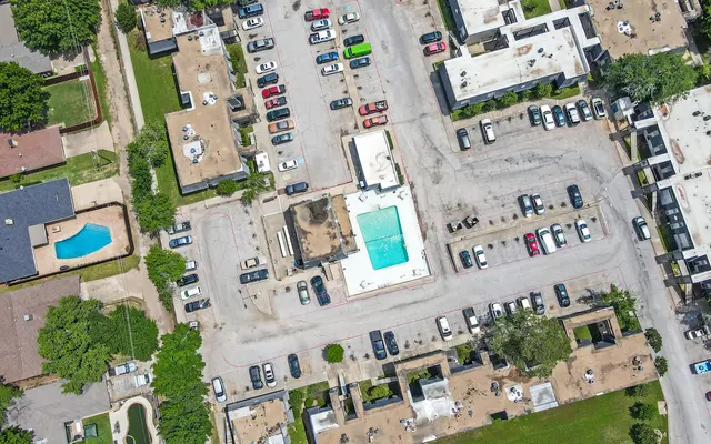 An aerial view of a residential area featuring a swimming pool, parking lot with numerous cars, and surrounding landscaped areas with trees.