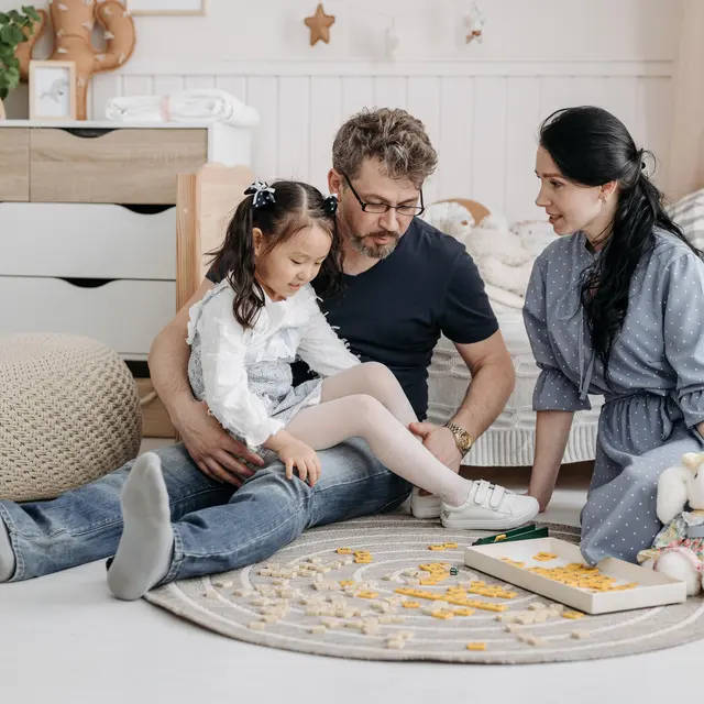 A family sitting on a soft rug playing a board game together. A father is helping his daughter while the mother watches and smiles, surrounded by a cozy living space.