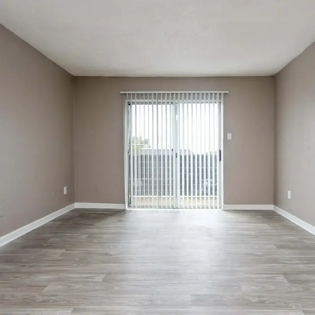 A spacious empty living room with a light brown wall and wooden flooring. There is a door on the left side and a sliding glass door with vertical blinds in the middle, leading outside.