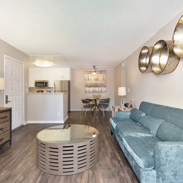 A modern living room featuring a light blue couch, wooden dresser, and decorative wall art. The flooring is wooden, and there are metallic wall decorations. A round coffee table sits in front of the couch, and there's a glimpse of a kitchen in the background.