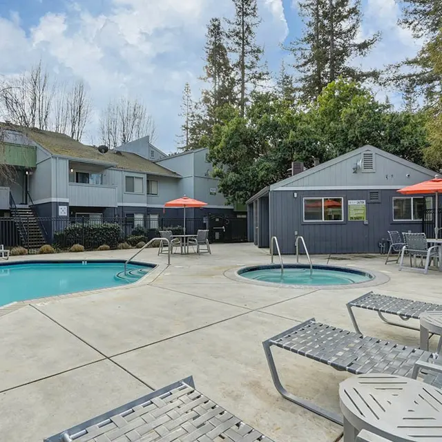 An outdoor pool area at an apartment complex, featuring a swimming pool, hot tub, and lounge chairs surrounded by greenery and buildings.