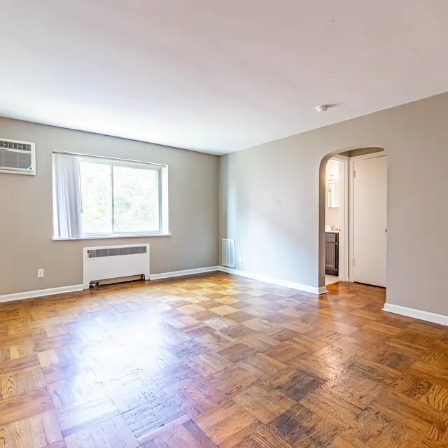 A spacious living room featuring a wooden floor, light-colored walls, and a large window allowing natural light to enter. There is an air conditioning unit mounted on the wall and a doorway leading to another room or kitchen area.