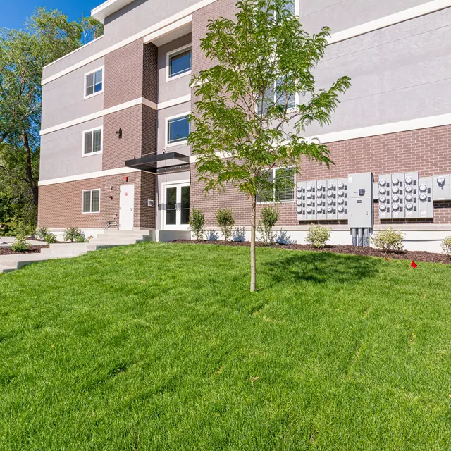 A modern apartment building with a landscaped lawn and trees in front. It features a three-story design with a combination of brick and gray exterior. Mailboxes are placed on the wall beside the entrance.