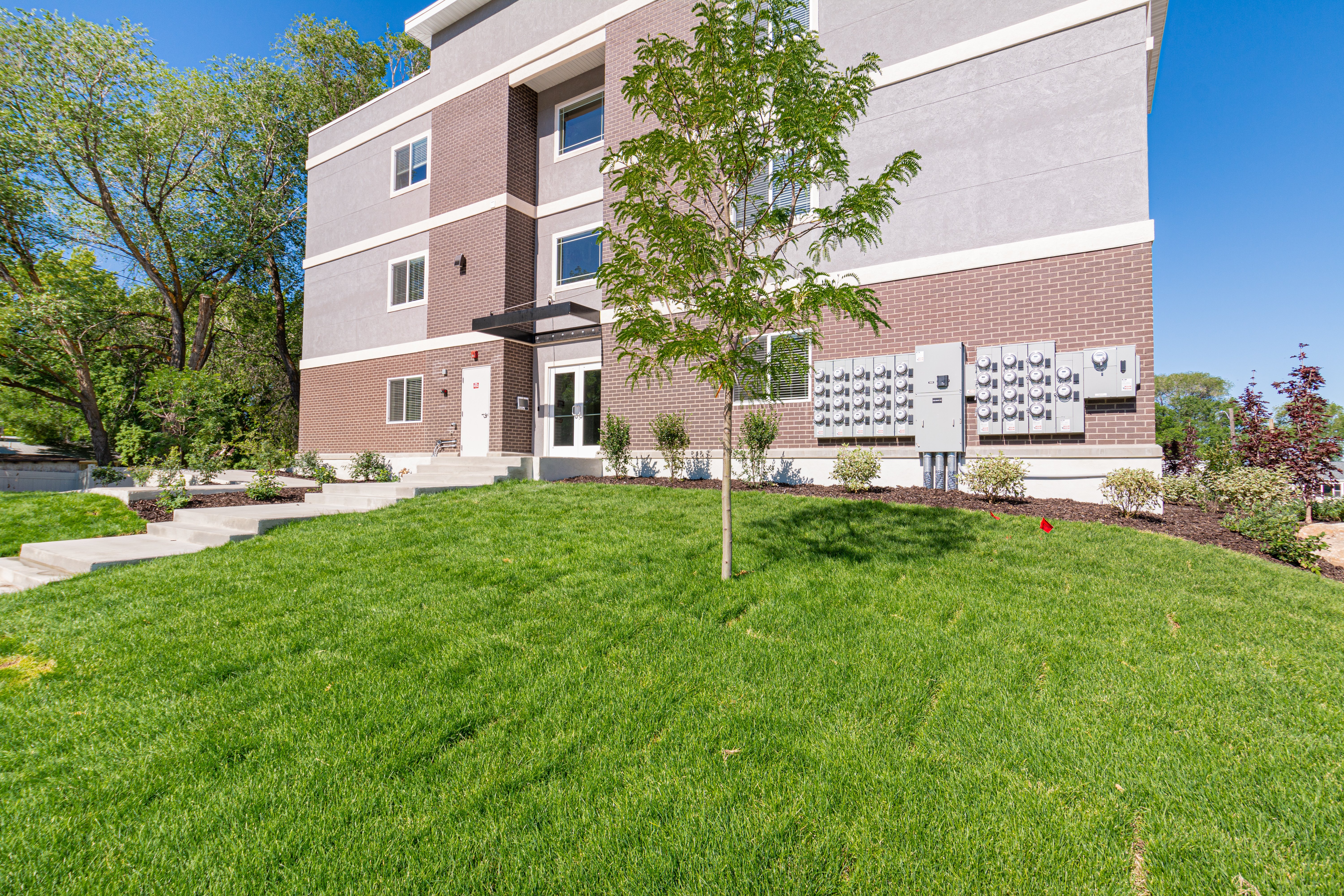 Modern Apartment Building Exterior A modern apartment building with a landscaped lawn and trees in front. It features a three-story design with a combination of brick and gray exterior. Mailboxes are placed on the wall beside the entrance.