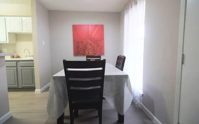 A cozy dining area featuring a wooden table with a white tablecloth, and black chairs. A large piece of red flower artwork hangs on the wall, and there are curtains letting in natural light from a window.