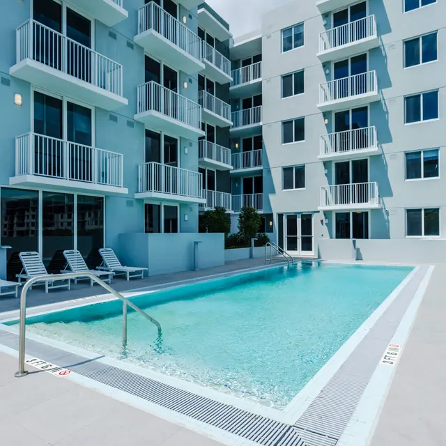A swimming pool with clear water surrounded by a modern apartment building. The pool area has lounge chairs neatly arranged on one side, and the building features balconies and large windows.