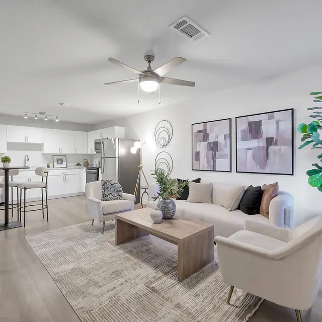 A modern living room featuring a white sofa with colorful pillows, a wooden coffee table, and a large plant. The space has grey walls and is decorated with abstract artwork. A kitchen area is visible in the background with white cabinets and stainless steel appliances.