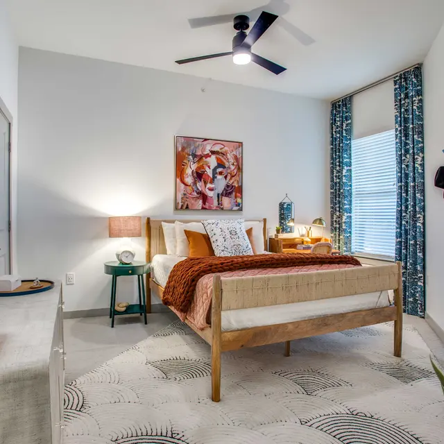 A modern bedroom featuring a wooden bed with a brown throw blanket and decorative pillows. There are two bedside tables with lamps, a circular mirror on the wall, artwork above the bed, and a sunlight streaming through the window with blue curtains. The floor is covered by a patterned rug and there is a plant in the corner.