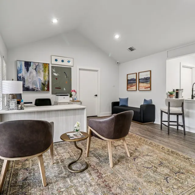 A stylish office interior featuring a reception desk, two modern chairs, and an open layout with a kitchen area in the background. The decor includes abstract artwork and a warm area rug.