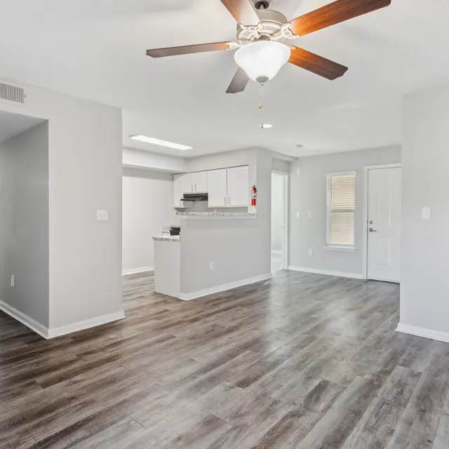 A modern, open living space with wood flooring, ceiling fan, and view of a kitchen and bathroom area in light colors.