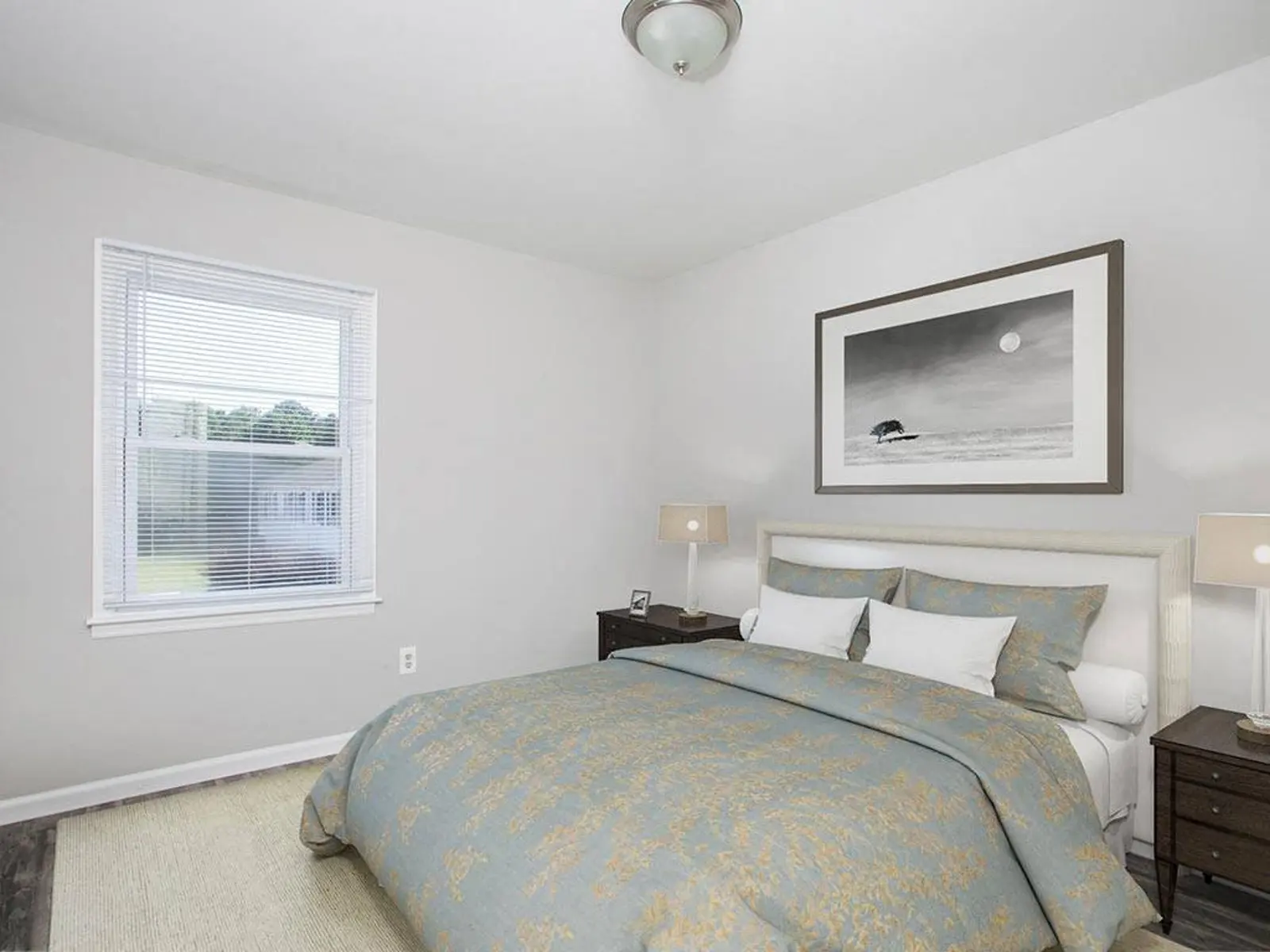 A cozy bedroom featuring a queen-sized bed with a blue and gold comforter, two bedside lamps, and a large framed black and white artwork on the wall. A window allows natural light to fill the room, with a view of the outdoors.