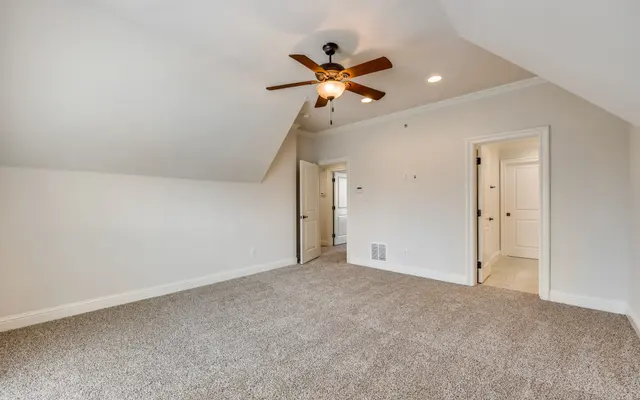 Empty Attic Room An empty attic room with a sloped ceiling, carpeted floor, ceiling fan, and a doorway leading to a small bathroom.
