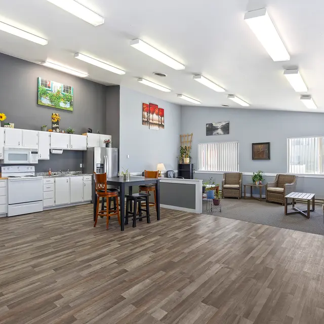 Community Room with Kitchen and Seating Area A spacious community room featuring a kitchen area and seating area. The kitchen has white cabinets and appliances, and there are tables and chairs set up for dining or activities. The seating area has comfortable chairs and a coffee table, with large windows allowing natural light to enter. The walls are adorned with pictures and decor, creating a welcoming atmosphere.