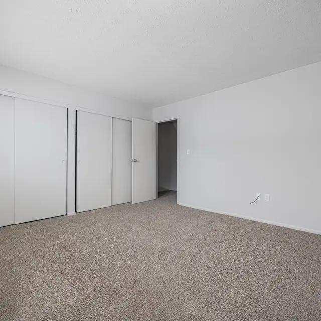 A spacious, empty room with beige carpet and light gray walls. There are two white closet doors on the left and an open doorway on the right leading to another room.