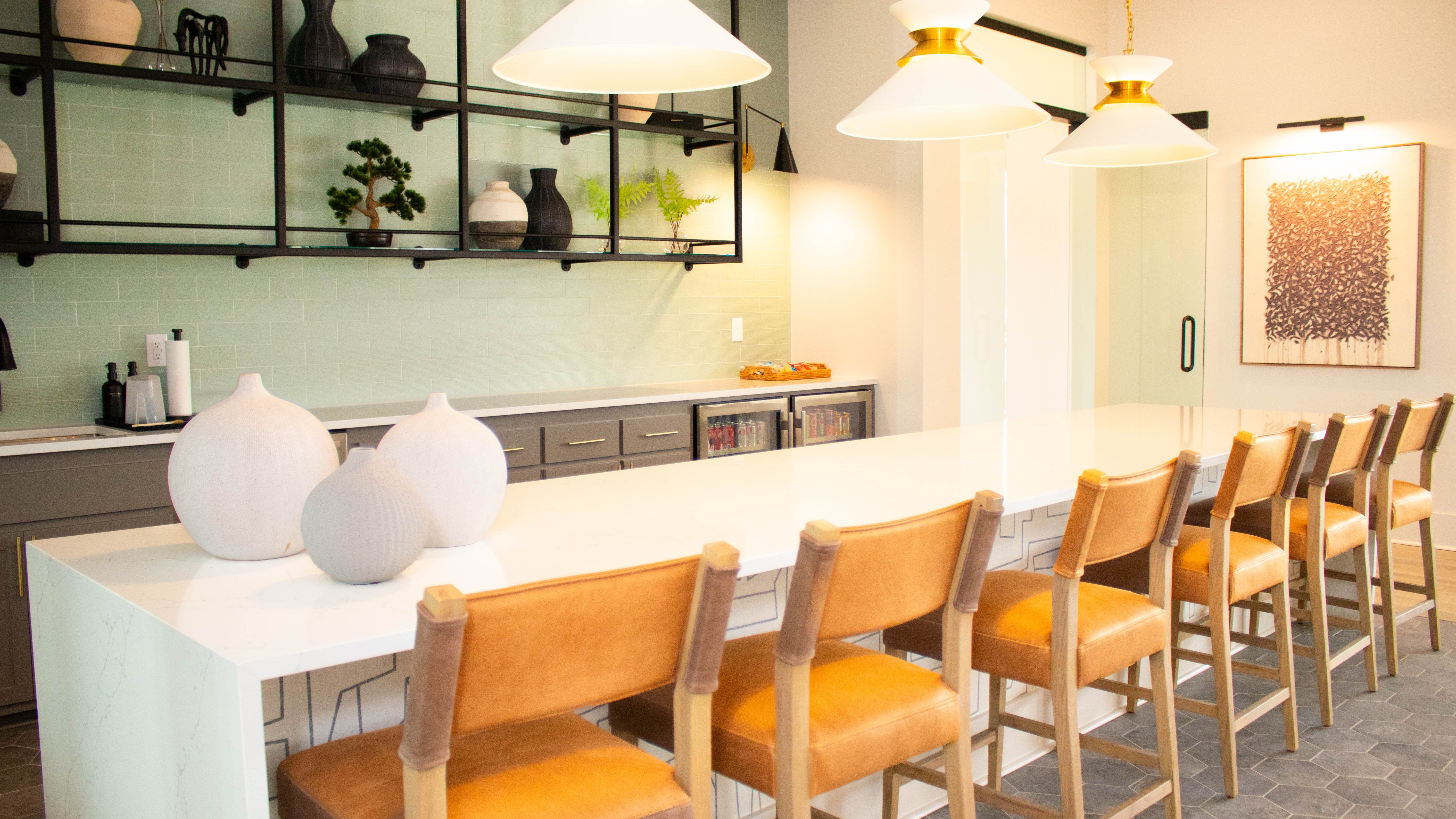 Modern Community Kitchen Space A modern kitchen area featuring a long white countertop with bar seating, pendant lights hanging above, and greenery on the shelves.