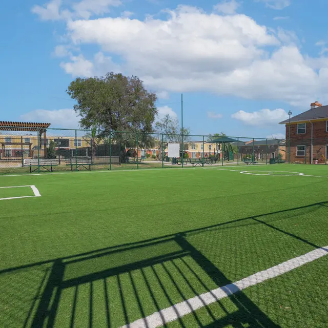 A well-maintained soccer field with a bright green artificial turf. In the background, there are apartment buildings and a tree, with a clear blue sky and fluffy white clouds overhead. A goal post is visible in the foreground.