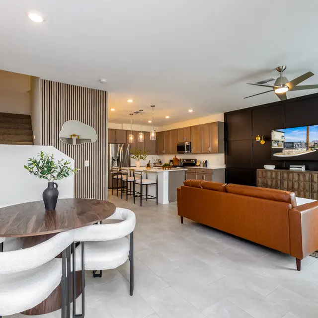 A stylish modern living area featuring a brown sofa, green armchair, dining table, and a kitchen in the background. The decor includes a mixture of light and dark tones with a mounted television on the wall.