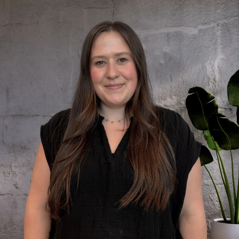 A young woman with long brown hair, smiling in front of a textured wall with a green plant beside her.