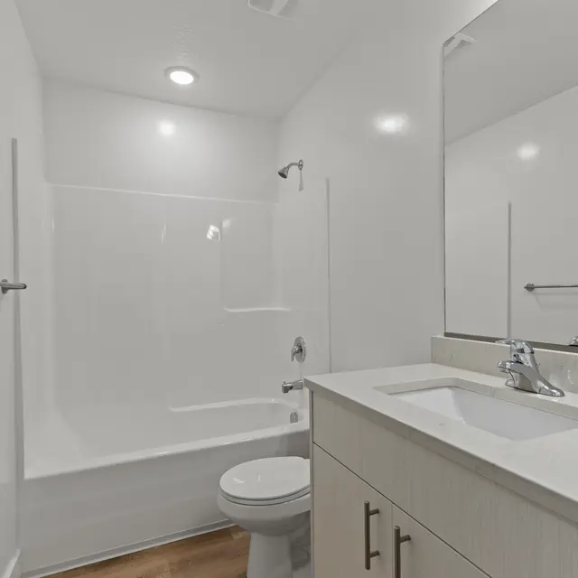 A modern bathroom featuring a white tub and shower, a toilet, and a double sink vanity. The walls and fixtures are bright and clean, with wooden flooring.