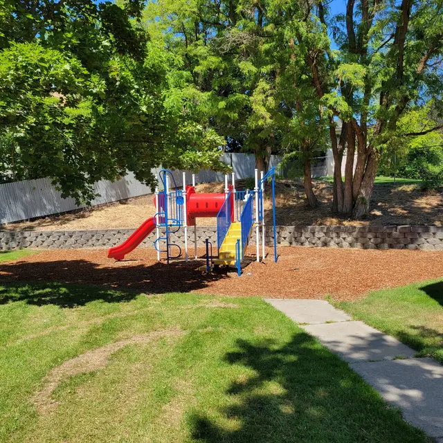 Colorful Playground with Slide A vibrant playground featuring a slide and climbing structure, situated in a grassy area with trees and a clear blue sky in the background.