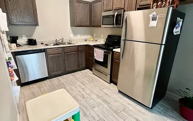 A contemporary kitchen featuring wooden cabinets, stainless steel appliances, and tile flooring. There's a small table with colorful stools in the foreground.