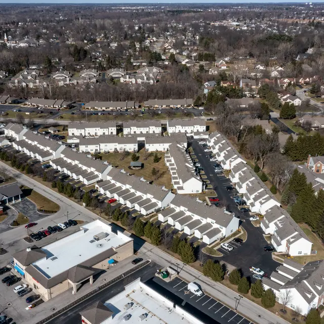 An aerial view of a residential area featuring townhouses, parking spaces, and some commercial buildings nearby. The landscape includes green trees and roads, with a sprawling suburban layout.