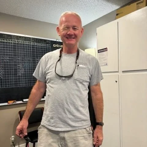 Cheerful Man in Gray Shirt A smiling man wearing a gray t-shirt and shorts stands with his arms outstretched in an indoor setting, with a blackboard in the background.