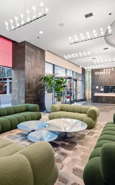 A modern lobby interior featuring elegant design elements. The space includes green, rounded sofas arranged in a circular pattern around a glass coffee table. The background showcases large windows with city views and bright lighting from pendant fixtures above. A reception area is visible in the back, adding a warm ambiance to the setting.