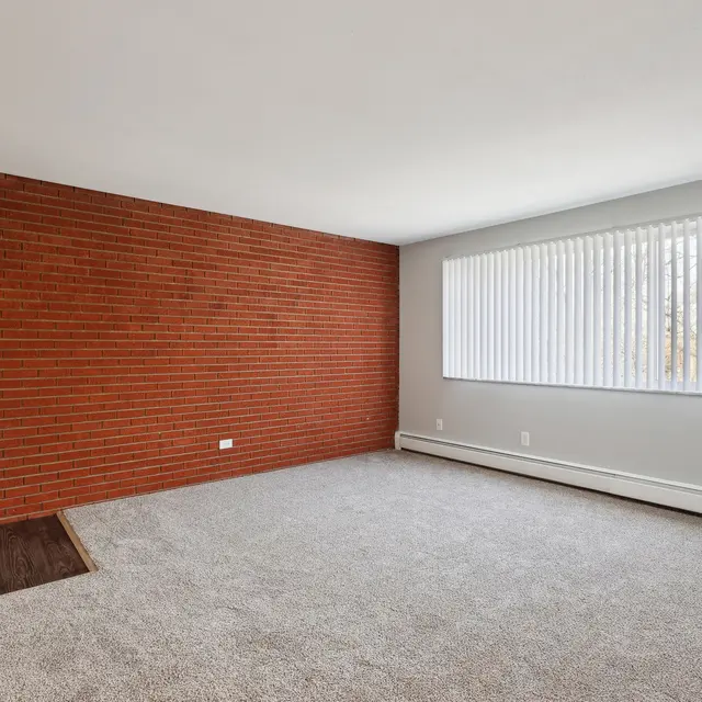A spacious empty living room featuring a textured brick wall on the left side, light-colored walls, and a large window with vertical blinds allowing natural light. The floor is covered with a light-colored carpet, and there's a small patch of flooring exposed.