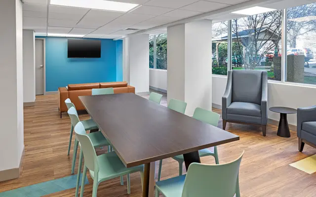 A modern office space featuring a rectangular table surrounded by colorful chairs, a brown couch, and large windows allowing natural light in.