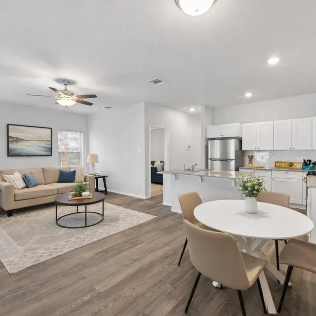 A spacious, modern living area with a beige couch, round coffee table, and dining area with a white circular table and chairs, featuring a contemporary kitchen in the background.